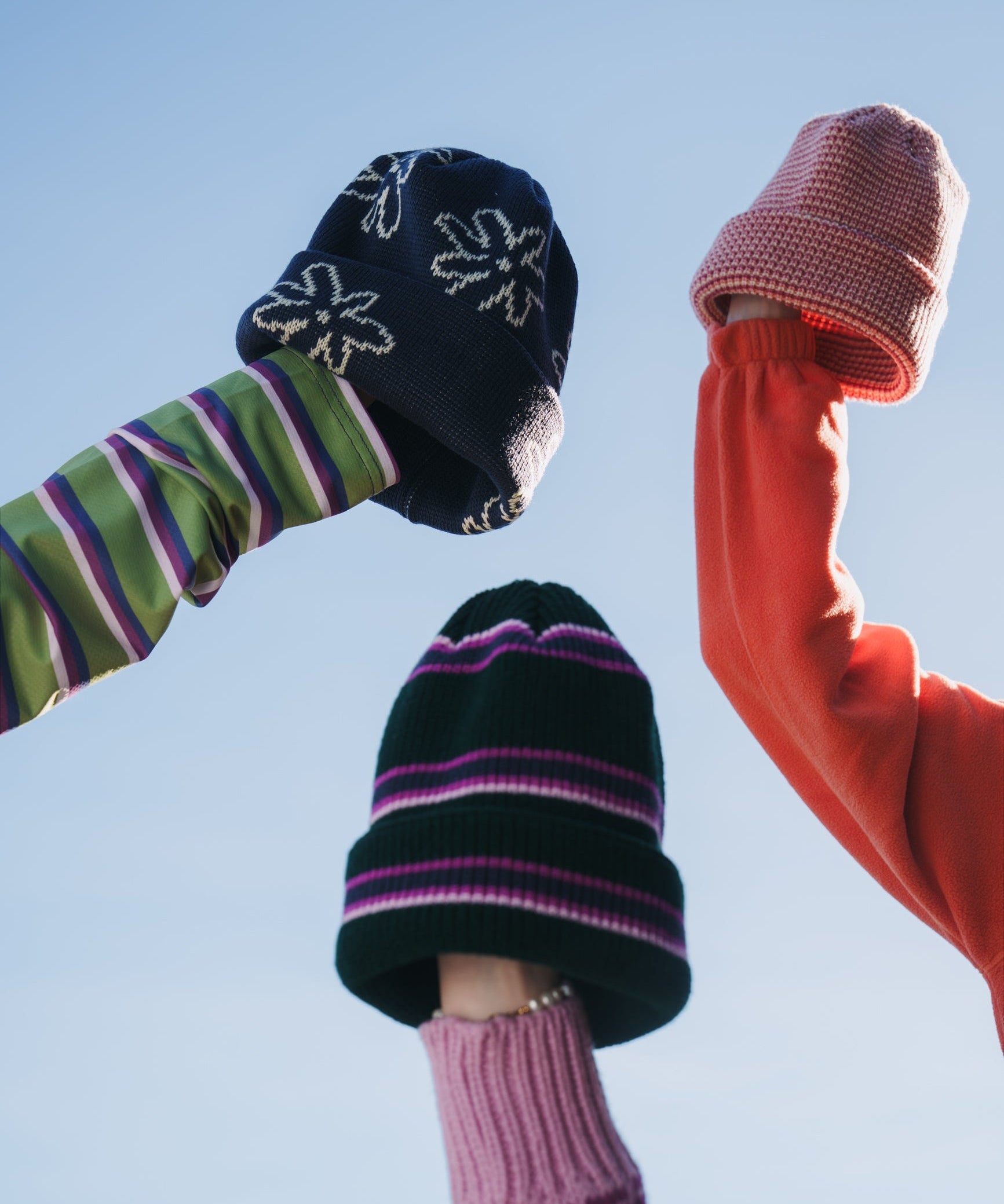 Three beanies held against a blue sky: navy floral, pink knit, and dark green with pink, purple and white stripes.