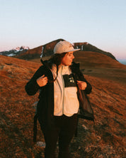 Women  standing on a mountain at sunset with a backpack wearing  beige off-white WOMEN fleece with green accent les Saisons