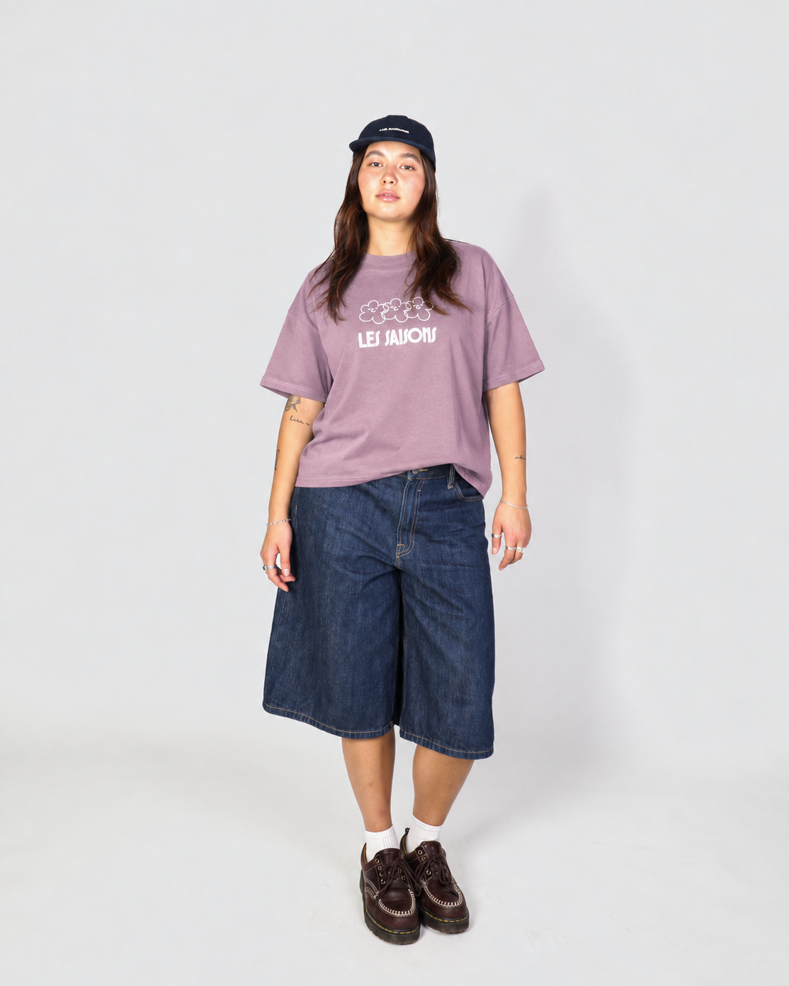 A model stands against a light gray studio background wearing a mauve-purple graphic t-shirt with 'LES Saisons' printed in white, dark-wash denim cropped shorts, white socks, brown loafers, and a black baseball cap.