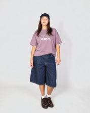 A model stands against a light gray studio background wearing a mauve-purple graphic t-shirt with 'LES Saisons' printed in white, dark-wash denim cropped shorts, white socks, brown loafers, and a black baseball cap.