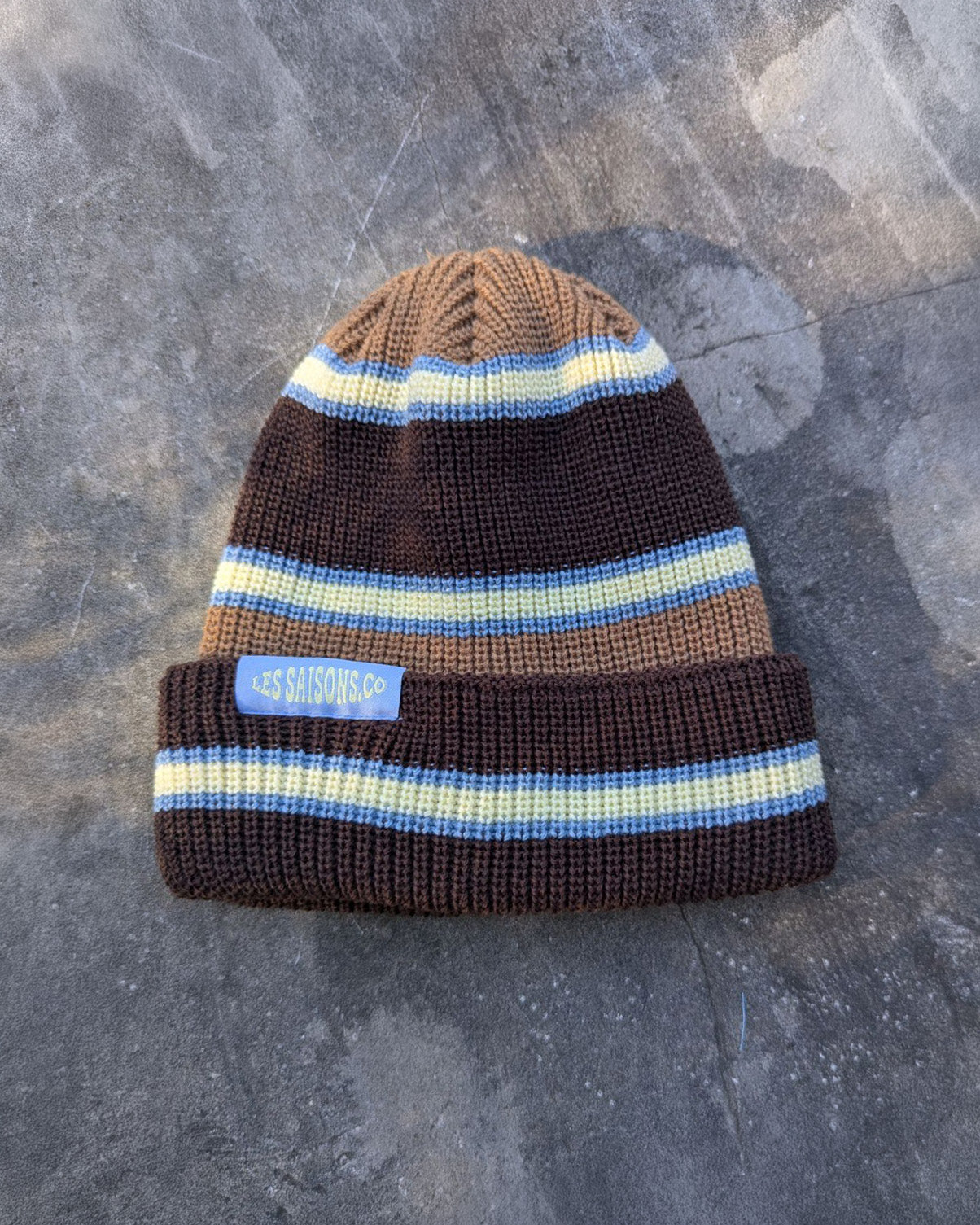 Brown and blue retro striped beanie with a blue les Saisons tag on a gray concrete surface, made in Canada.