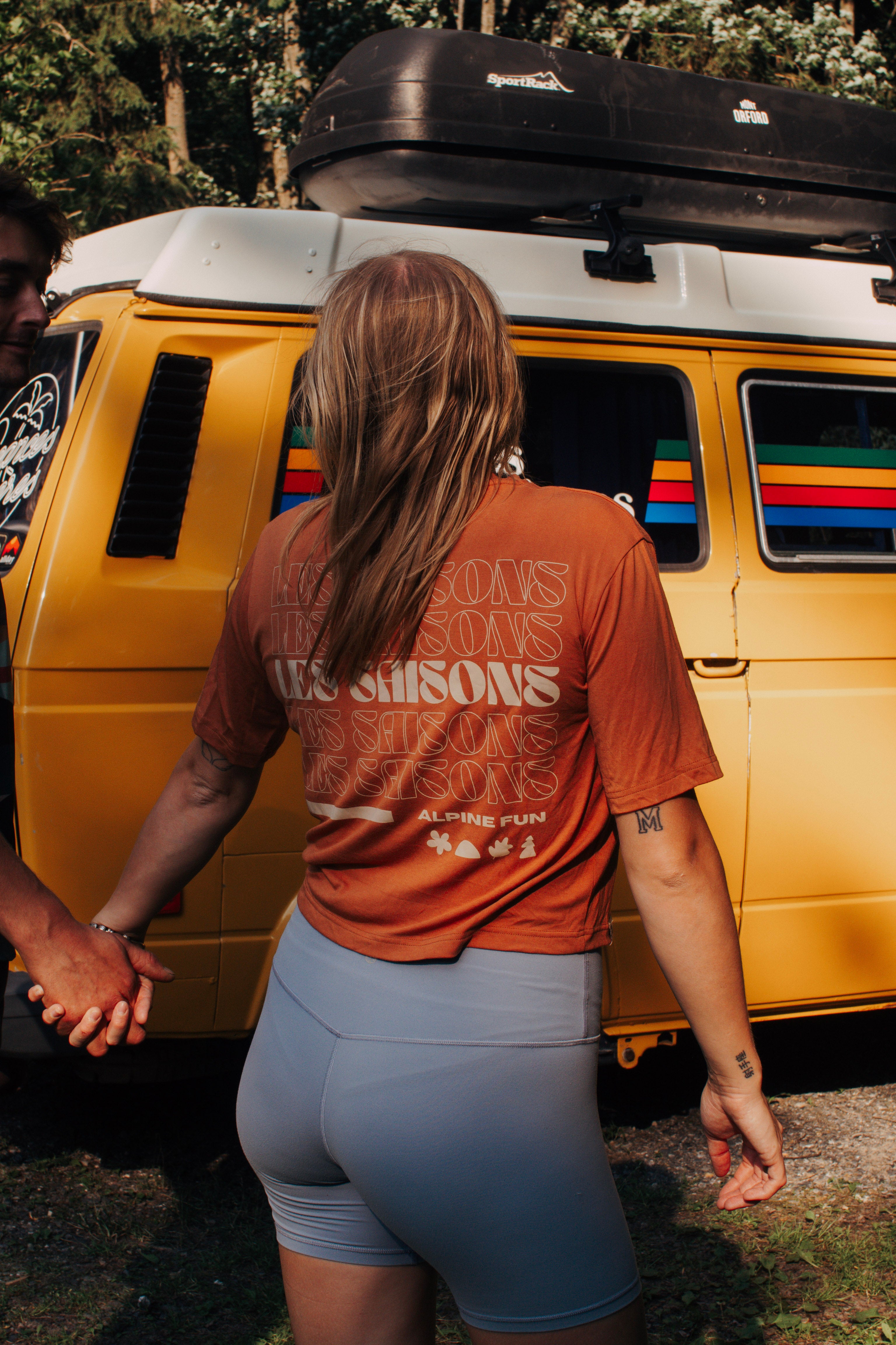 Person wearing a brown shirt with text standing next to a yellow van with a colorful stripe.