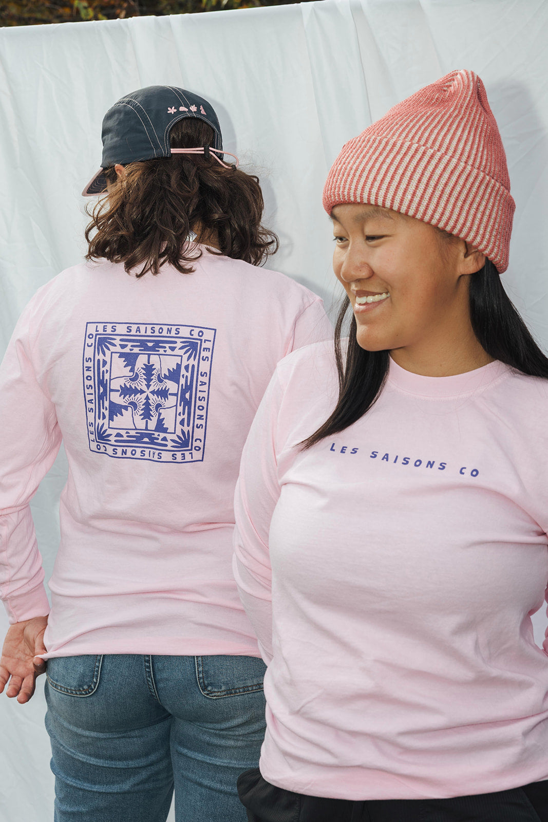 Two people wearing pink long-sleeve shirts with 'Les Saisons Co' branding, one with a graphic on the back.