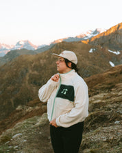 Women standing on a mountain with snow-capped peaks in the background wear the beige off-white WOMEN fleece with green accent les Saisons