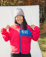 Person wearing a red and blue fleece jacket les Saisons with a colorful knit hat in an outdoor setting.