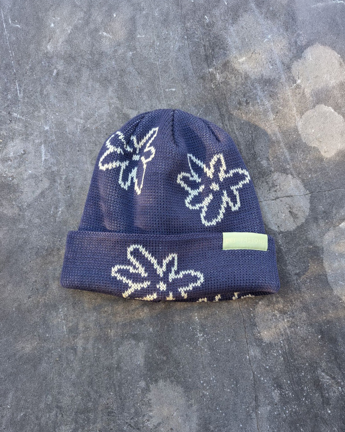 Navy beanie with white flowers with a green les Saisons tag on a gray concrete surface, made in Canada.