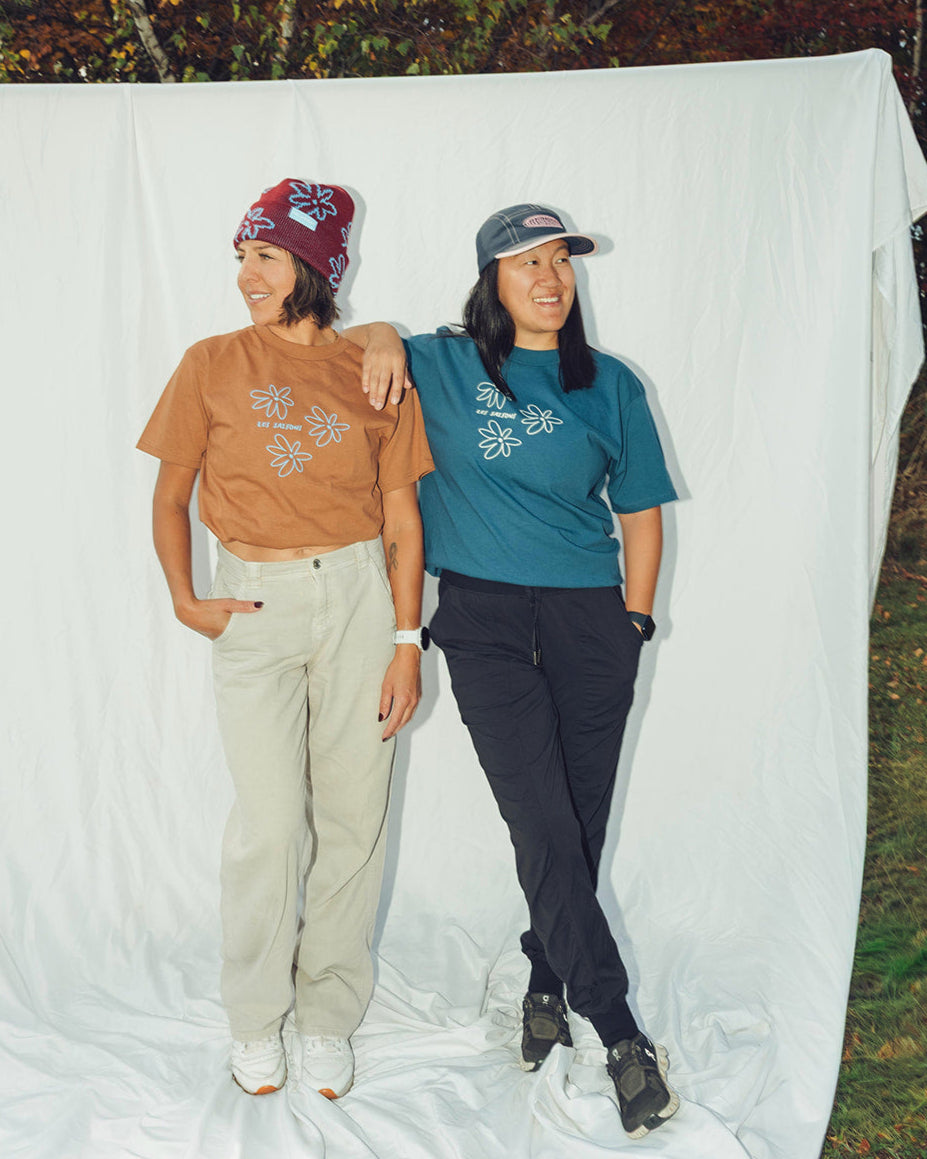 Two people standing in front of a white backdrop with trees in the background with flower Power Tshirts from les Saisons
