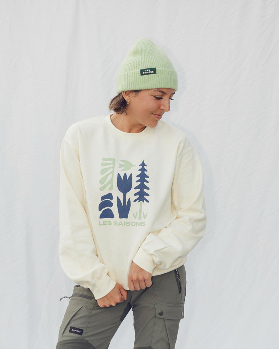Person wearing a white sweatshirt  unisex les Saisons with blue designs and a green beanie in front of a white background with trees in the background.