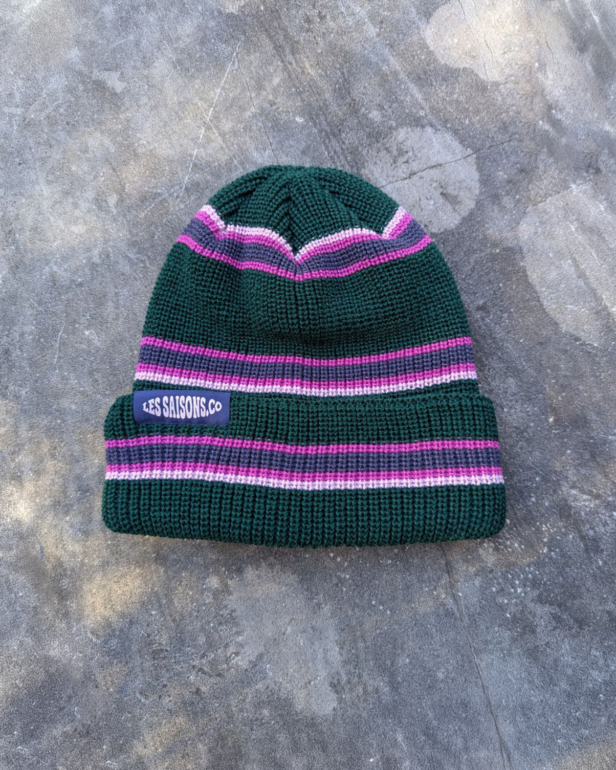 Green and pink retro striped beanie with a purple les Saisons tag on a gray concrete surface, made in Canada.