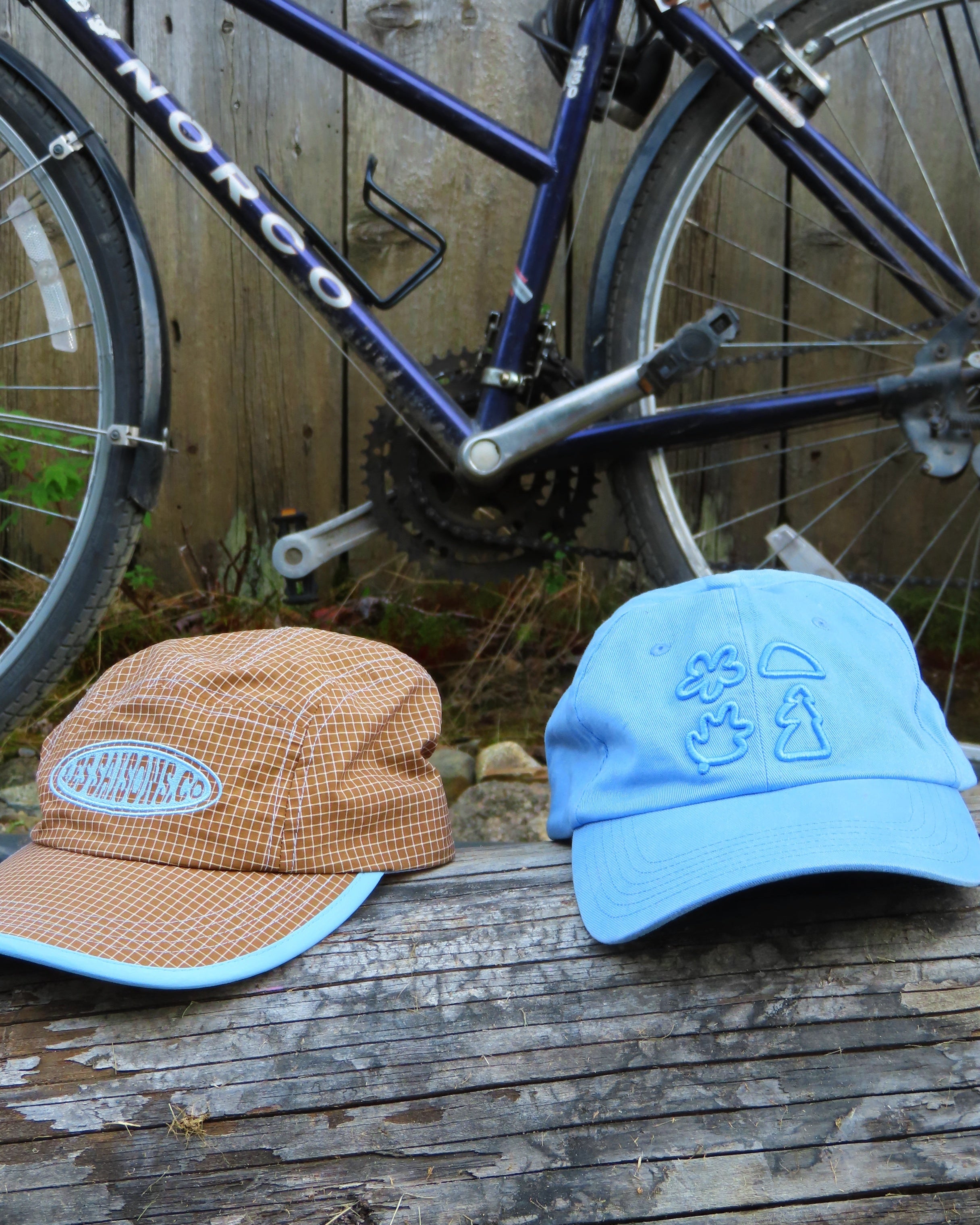 Two hats, one brown and one blue, on a wooden surface with a bicycle in the background.