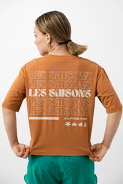 Person wearing an brown t-shirt with Les Saisons on the back, standing against a white background