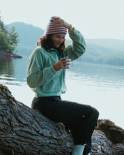 Person sitting on a log by a lake, wearing a light green jacket and striped beanie.
