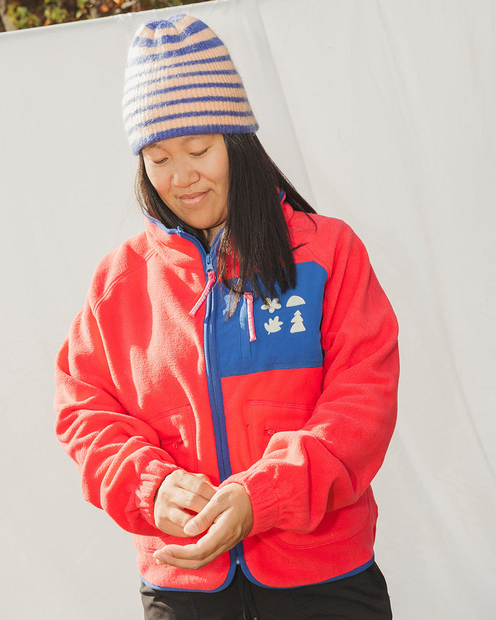 Person wearing a red jacket with a blue logo and a striped beanie in an outdoor setting.