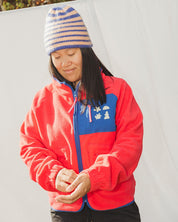 Person wearing a red jacket with a blue logo and a striped beanie in an outdoor setting.