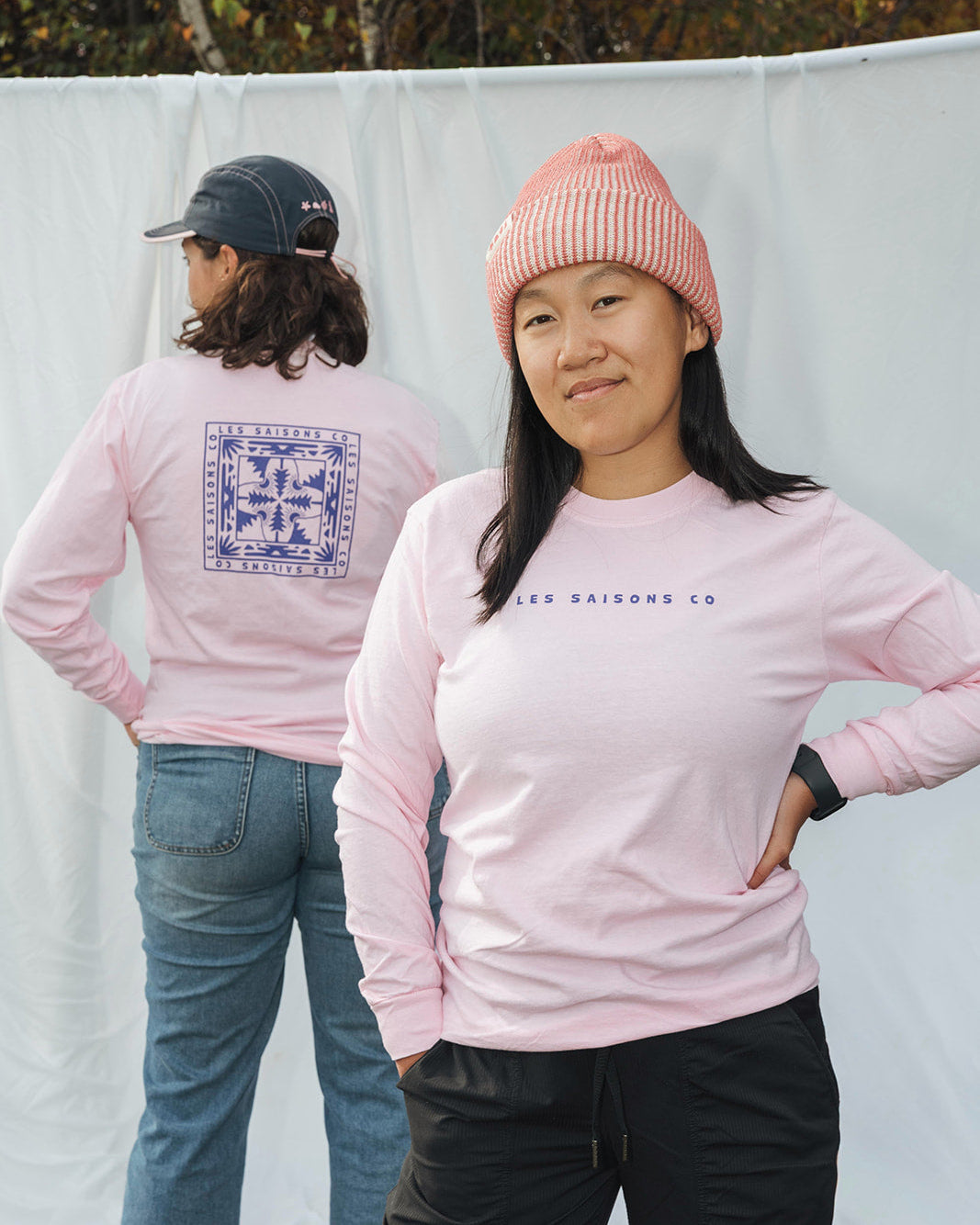Two people wearing pink  longsleeve sweatshirts with visible branding les Saisons against a white backdrop.