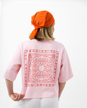 Person wearing a pink t-shirt with a red geometric pattern on the back, orange headscarf, and white pants against a white background.