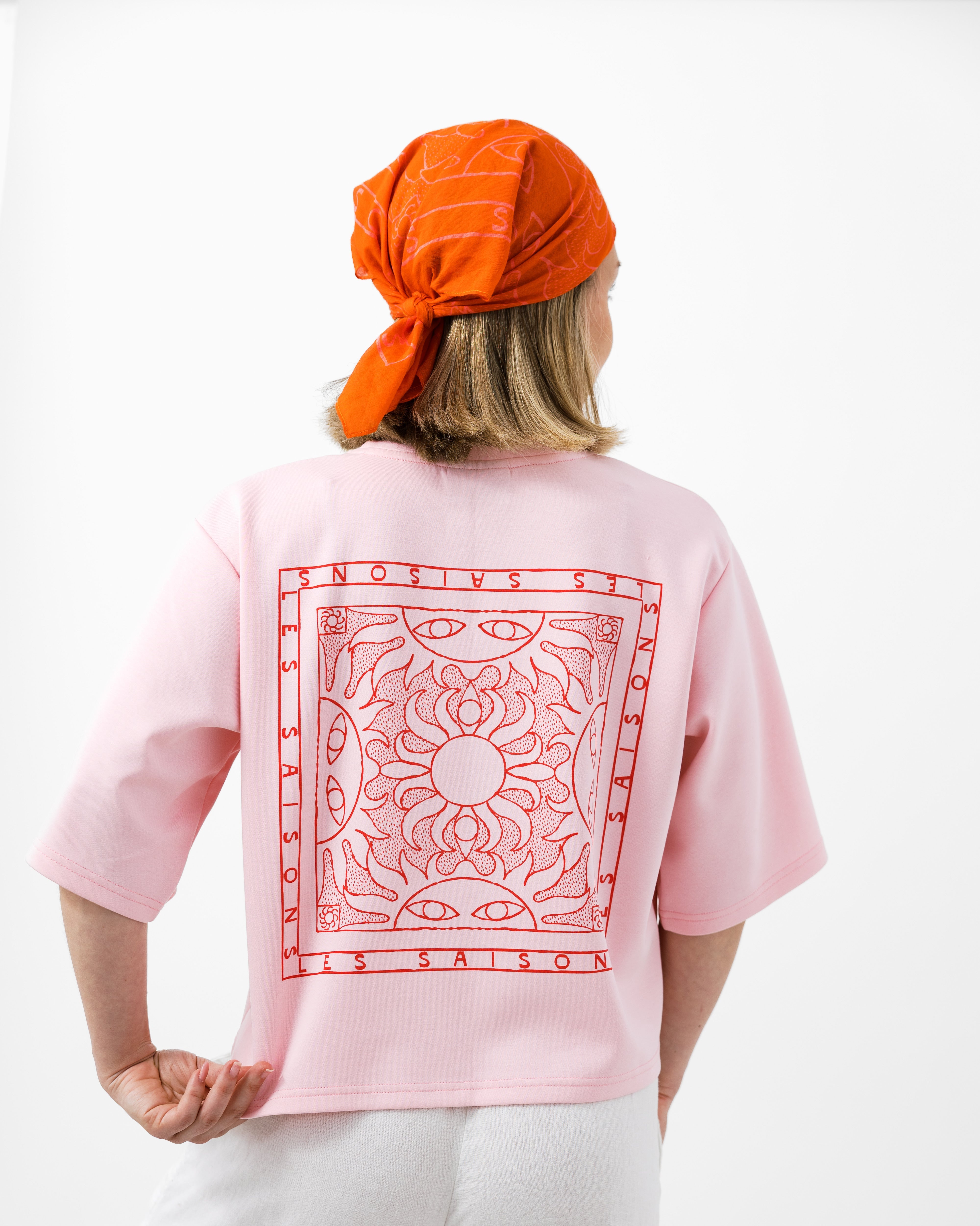Person wearing a pink t-shirt with a red geometric pattern on the back, orange headscarf, and white pants against a white background.