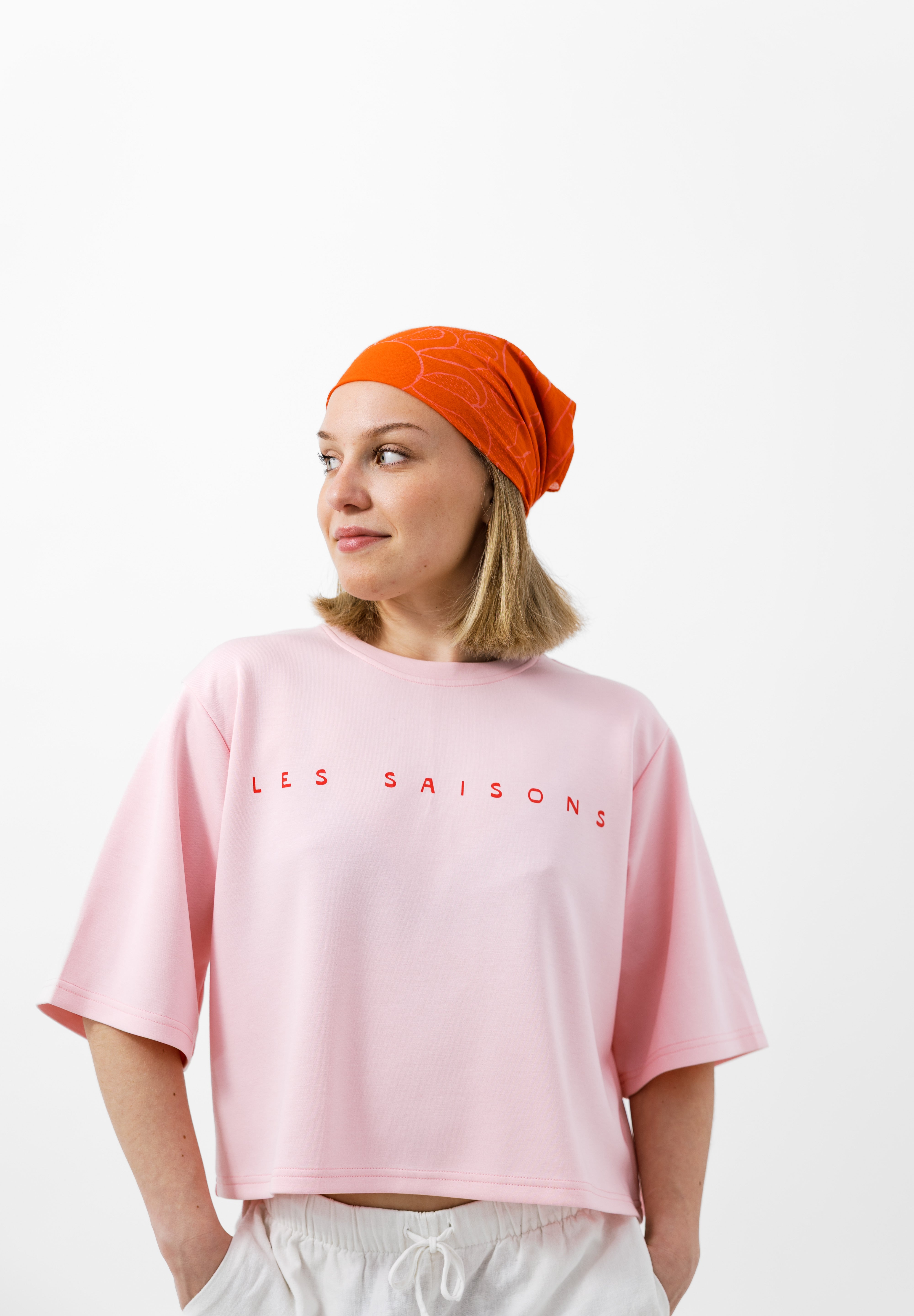 Person wearing a pink 'LES SAISONS' t-shirt and orange headscarf on a white background