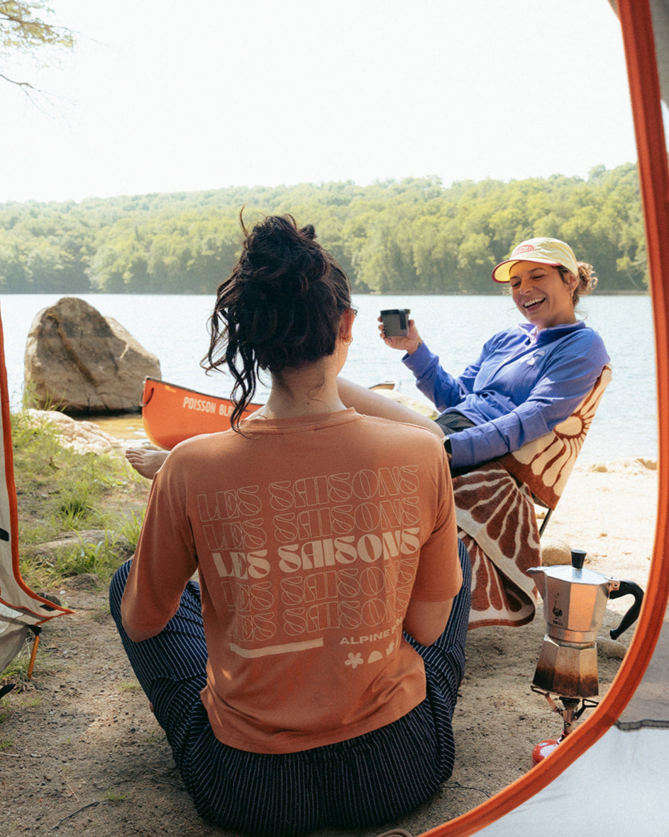 Two women sitting by a lake, one taking a photo, viewed from inside a tent.
