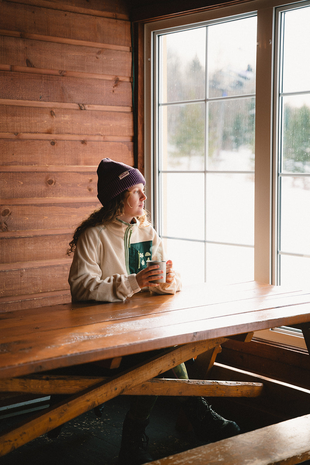 Girl sitting in a ski shack wearing Les Saisons beige fleece jacket drinking a hot cocoa