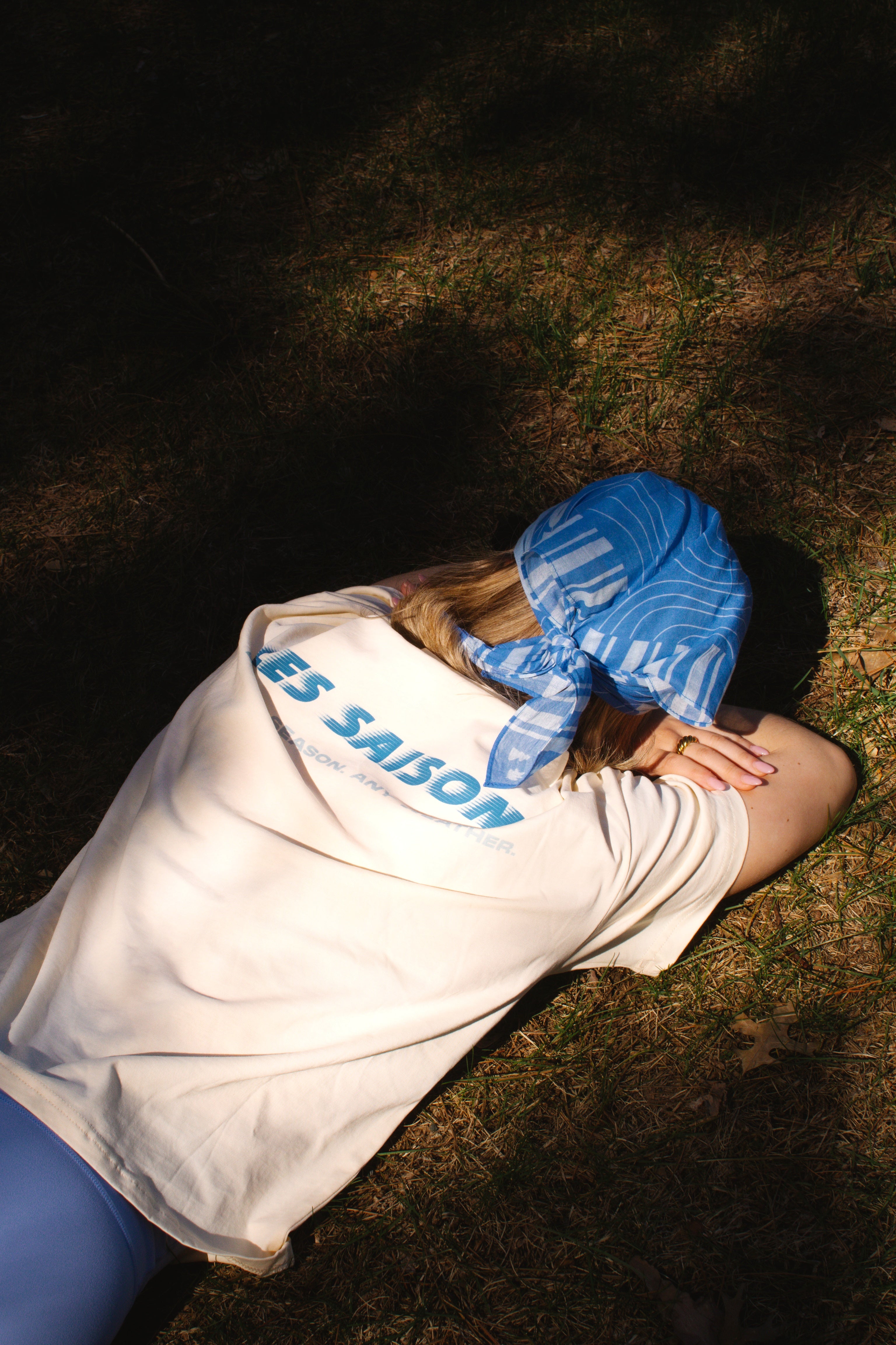 Person lying on grass wearing a butter colored tshirt with text and a blue bandana les saisons
