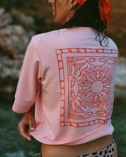 A woman wearing a light pink cropped T-shirt with a large red graphic print on the back, photographed outdoors with a blurred natural background.