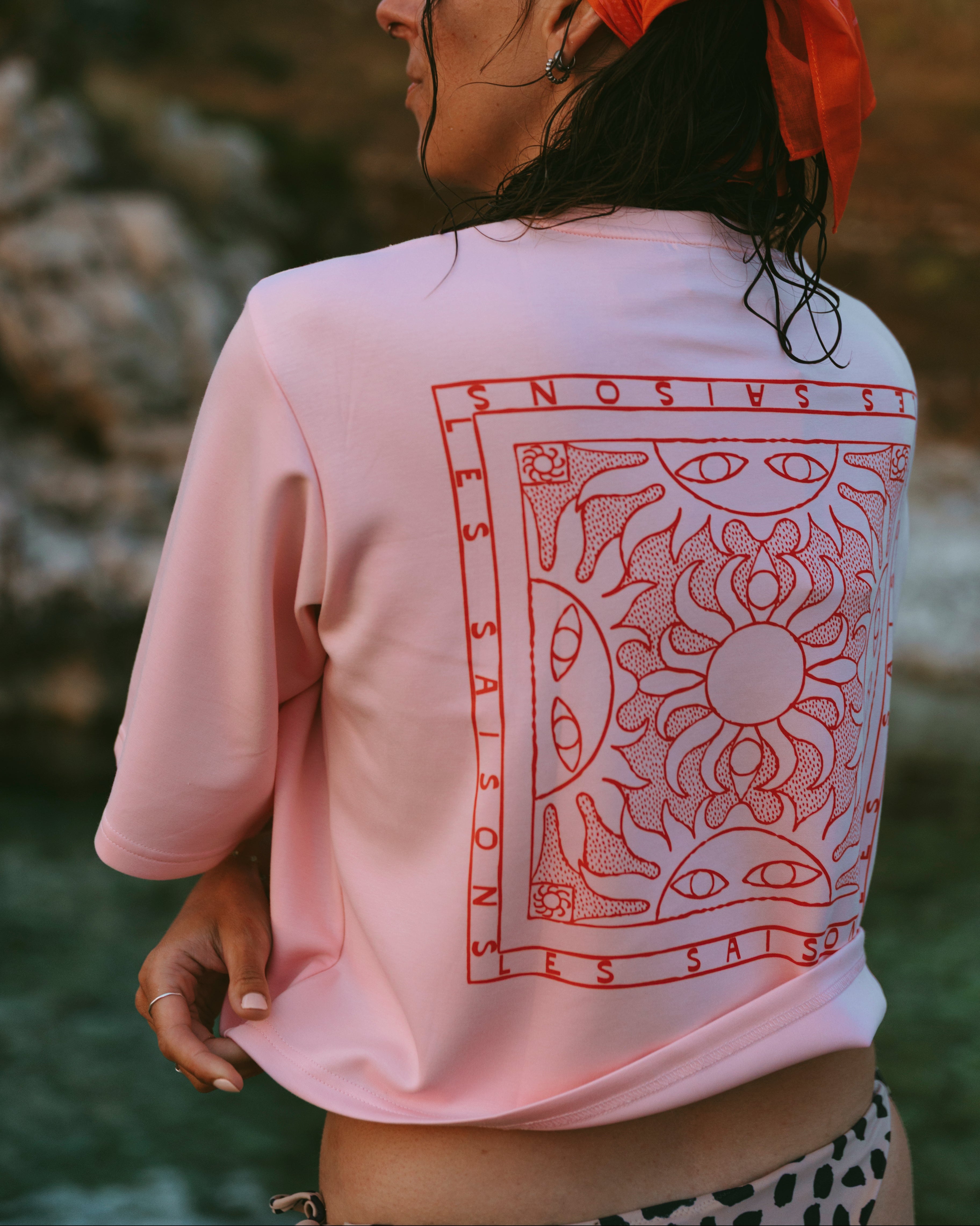 A woman wearing a light pink cropped T-shirt with a large red graphic print on the back, photographed outdoors with a blurred natural background.