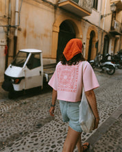 A woman wearing a light pink t-shirt with a red print on the back, a bright orange headscarf, denim shorts, and carrying a white shoulder bag.