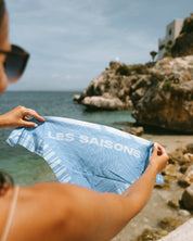 A person at a rocky beach holds up a light blue striped bandana with the words 'LES SAISONS' printed, with the sea and coastline blurred in the background.