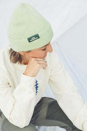 Person wearing a light green beanie with a logo, white hoodie, and gray pants on a light background