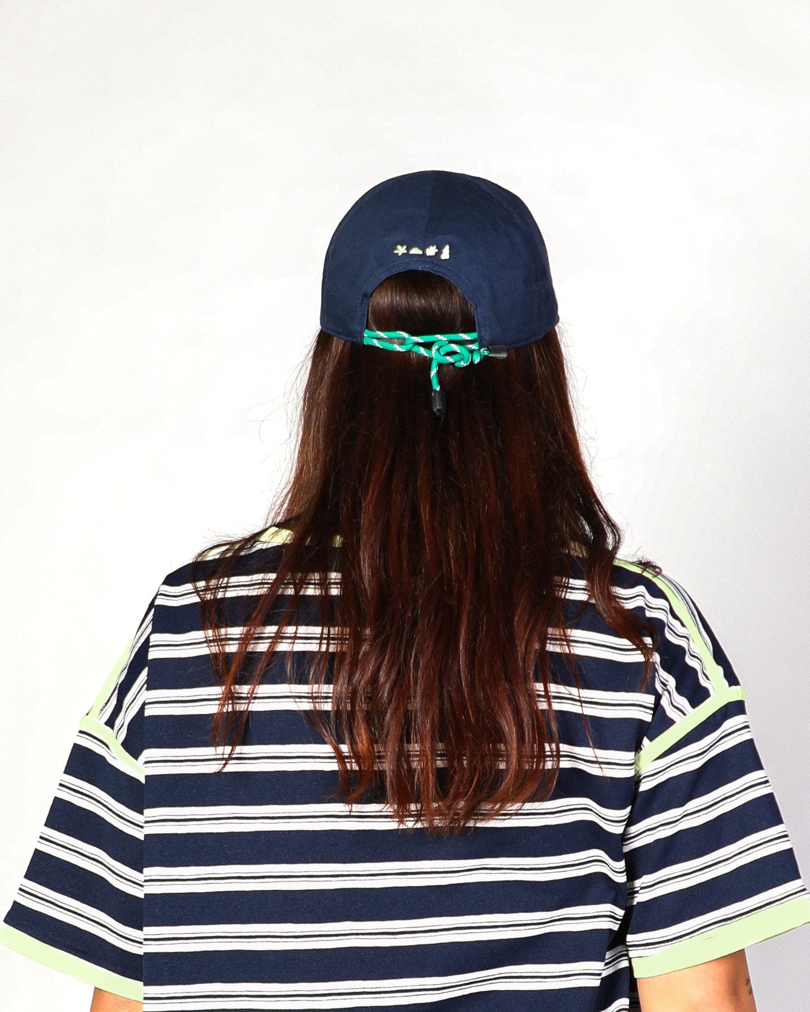 A person viewed from behind wearing a navy blue baseball cap with a green cord detail, a navy blue and white striped t-shirt with light green trim, and long dark brown hair against a plain white background.