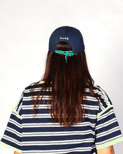 A person viewed from behind wearing a navy blue baseball cap with a green cord detail, a navy blue and white striped t-shirt with light green trim, and long dark brown hair against a plain white background.