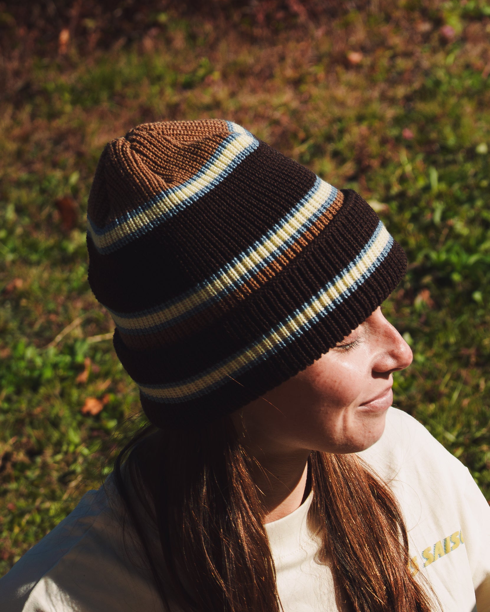 Person outdoors wearing a brown ribbed beanie with light blue and cream stripes and a folded cuff, shown in side profile.