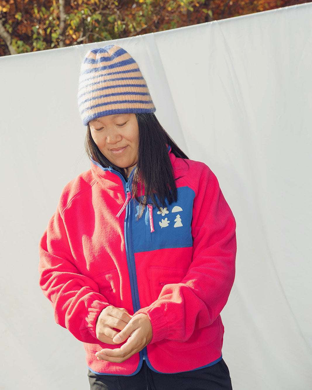 Person wearing a pink fleece jacket les Saisons with a blue zipper and colorful beanie in an outdoor setting.