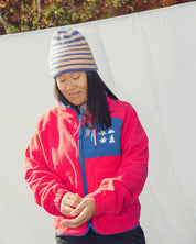 Person wearing a pink fleece jacket les Saisons with a blue zipper and colorful beanie in an outdoor setting.