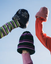 Three beanies held against a blue sky: navy floral, pink knit, and dark green with pink, purple and white stripes.