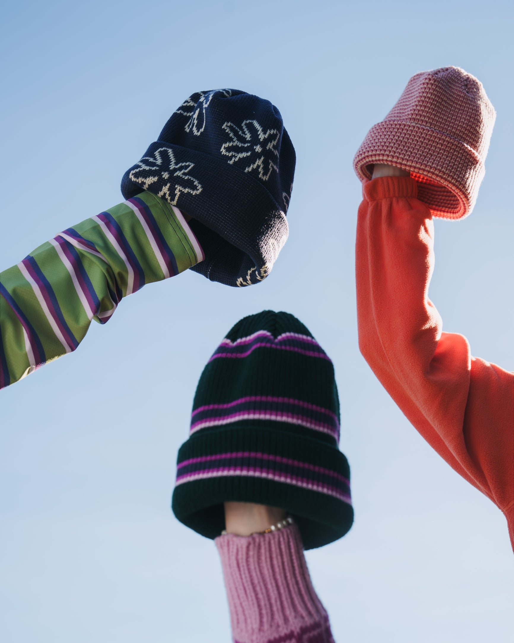 Three beanies held against a blue sky: navy floral, pink knit, and dark green with pink, purple and white stripes.