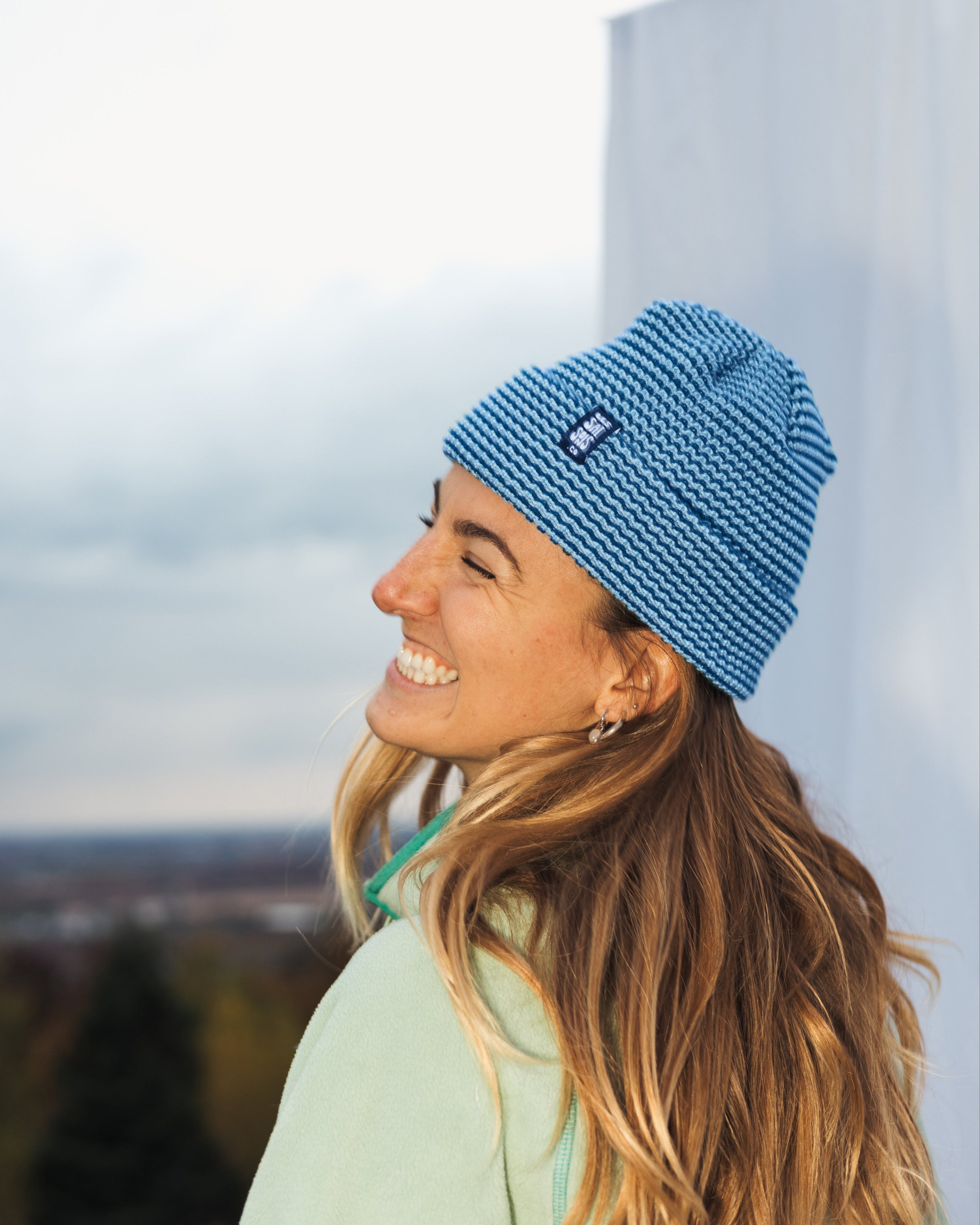 Woman wearing a blue knit beanie with a brand logo, standing against a light-colored wall with a blurred natural background.