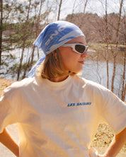 Outdoors person wearing a butter t-shirt with 'LES SAISONS' text, sunglasses, and a blue bandana for Running and hiking outdoors by a lake.