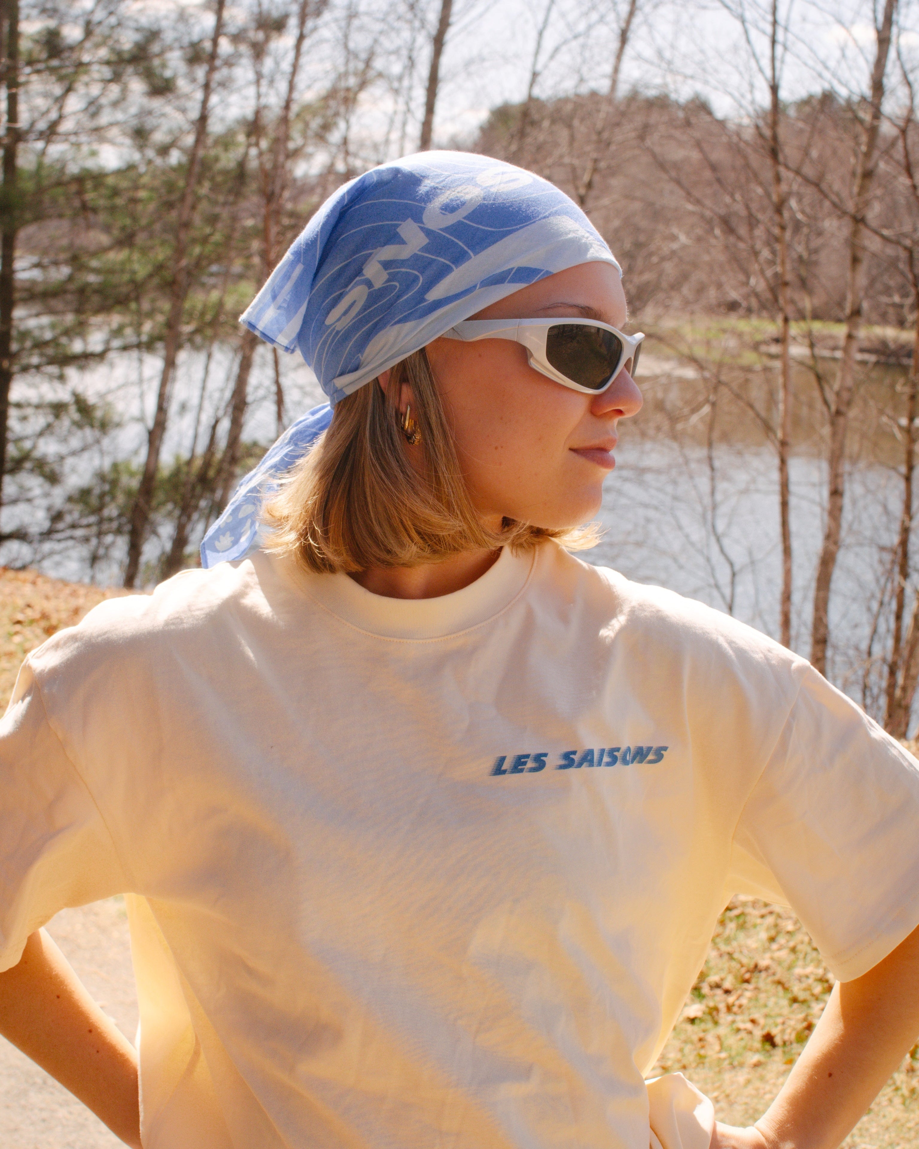 Outdoors person wearing a butter t-shirt with 'LES SAISONS' text, sunglasses, and a blue bandana for Running and hiking outdoors by a lake.