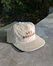 Beige corduroy cap with 'LES SAISONS' embroidery on a concrete surface with greenery in the background