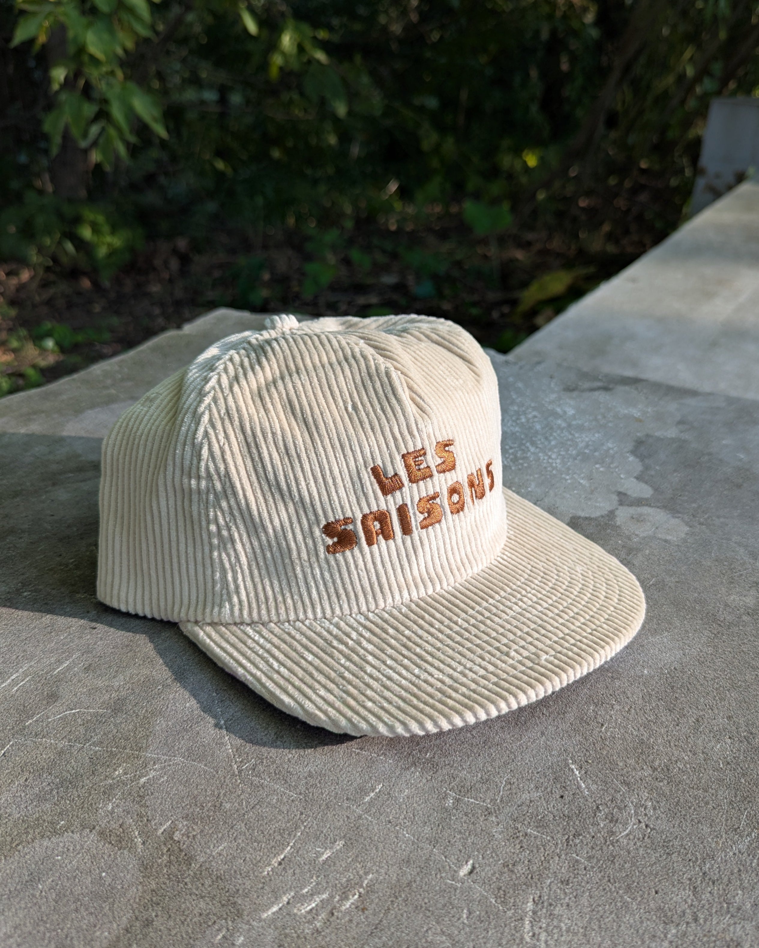Beige corduroy cap with 'LES SAISONS' embroidery on a concrete surface with greenery in the background