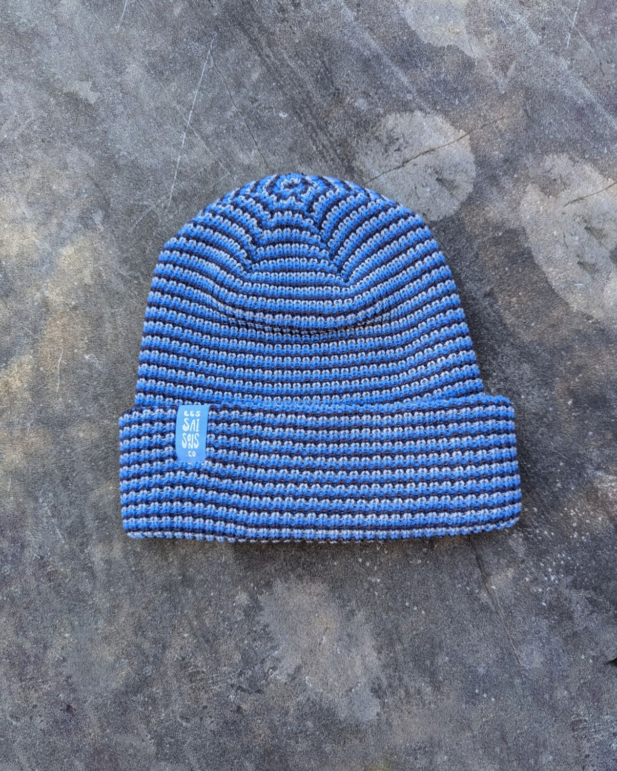 blue beanie with a blue les Saisons tag on a gray concrete surface, made in Canada.
