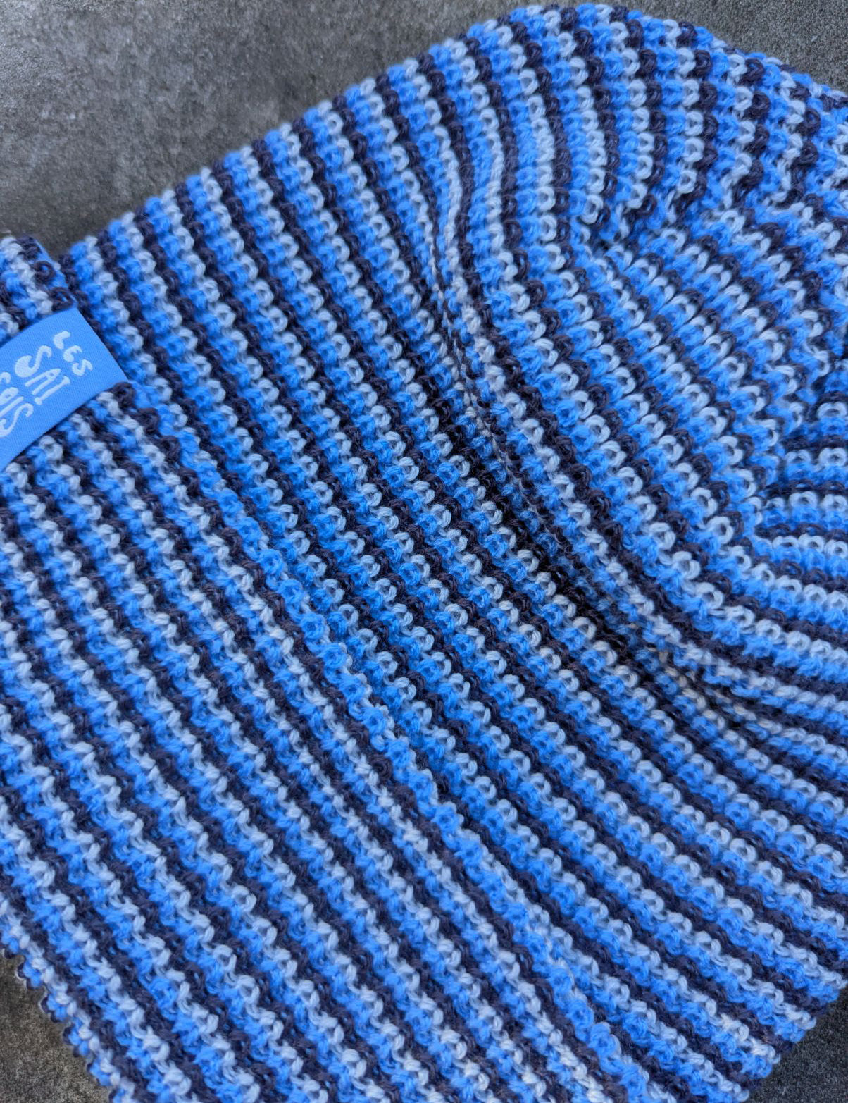 zoom on the blue beanie with a blue les Saisons tag on a gray concrete surface, made in Canada.