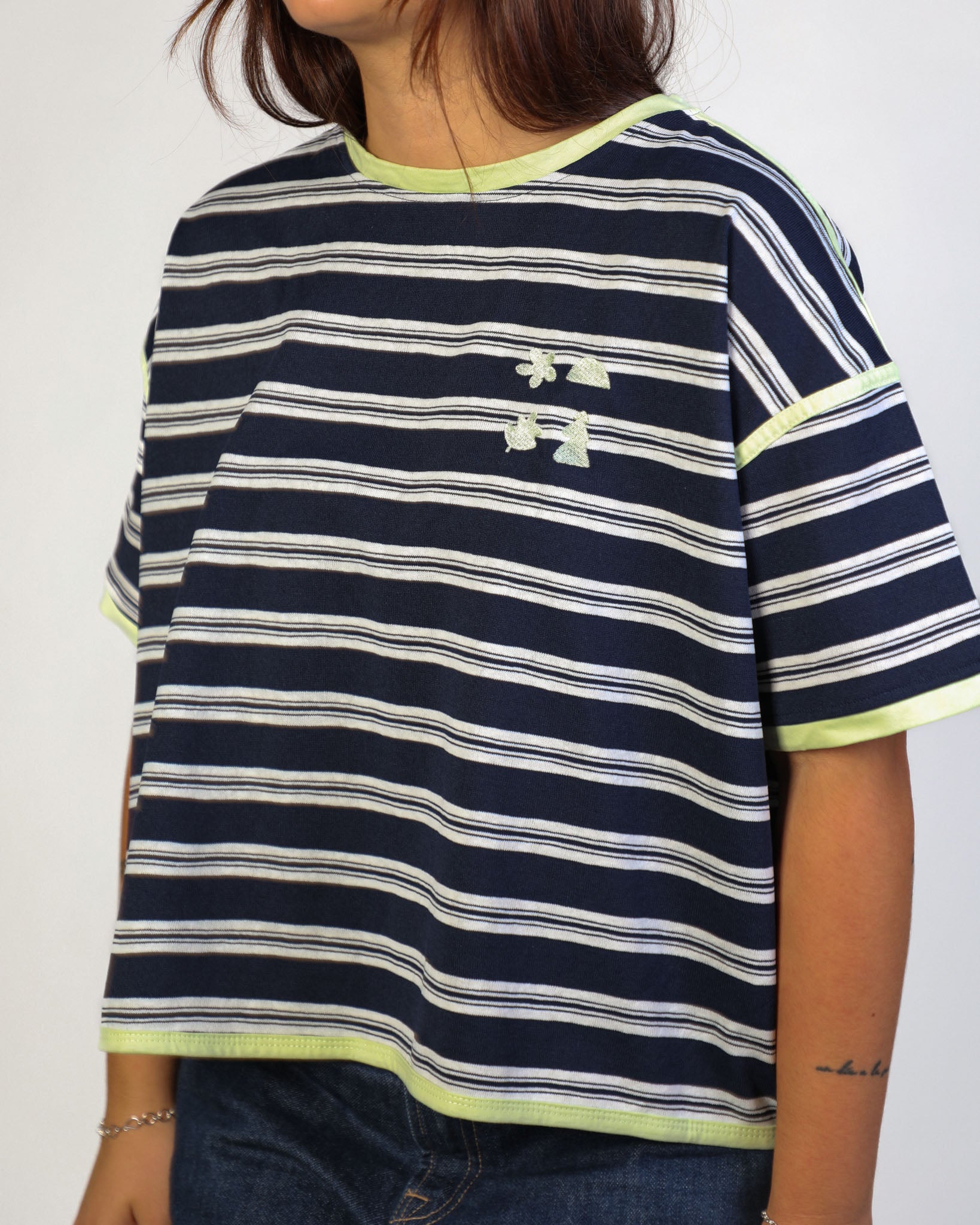 A person wearing a cropped short-sleeve t-shirt with navy blue, white, and lime green horizontal stripes, featuring a small white embroidered Under Armour logo on the chest.