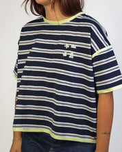 A person wearing a cropped short-sleeve t-shirt with navy blue, white, and lime green horizontal stripes, featuring a small white embroidered Under Armour logo on the chest.