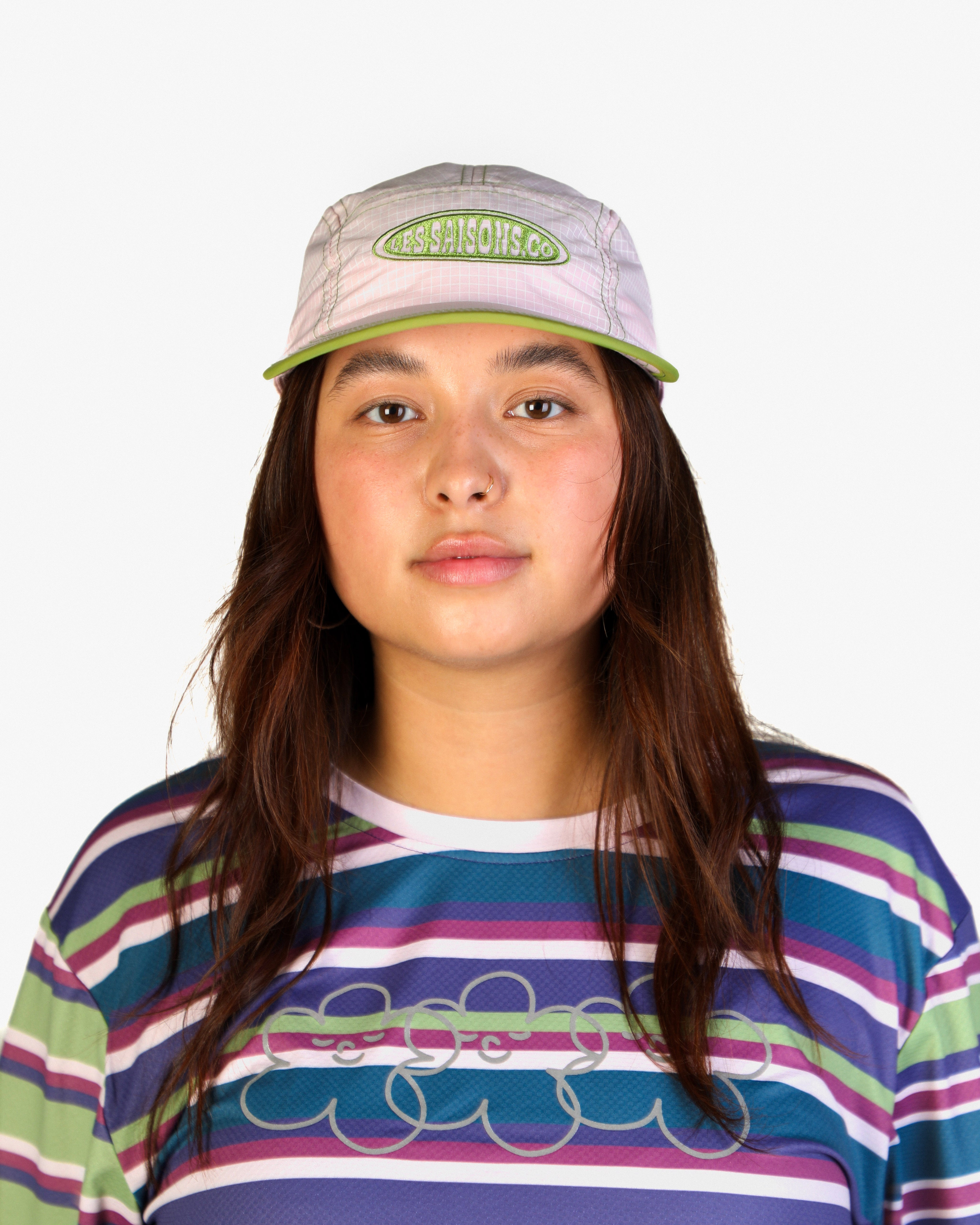 A young woman with long brown hair is shown from the chest up against a plain light gray background. She is wearing a light gray and green cap with the brand name 'Cruel Pigs' embroidered on the front, and a colorful horizontally striped t-shirt with a gray graphic.
