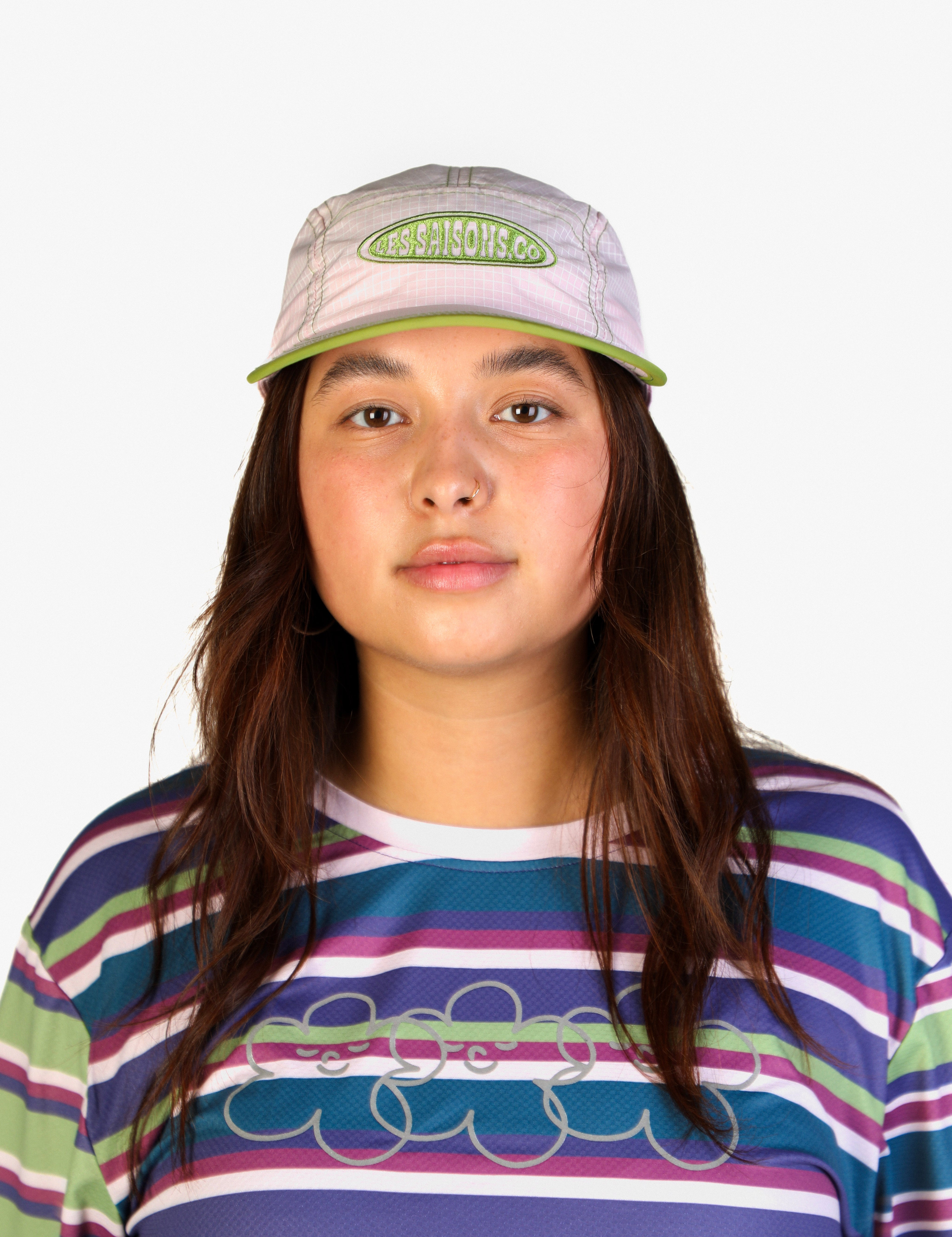 A young woman with long brown hair is shown from the chest up against a plain light gray background. She is wearing a light gray and green cap with the brand name 'Cruel Pigs' embroidered on the front, and a colorful horizontally striped t-shirt with a gray graphic.