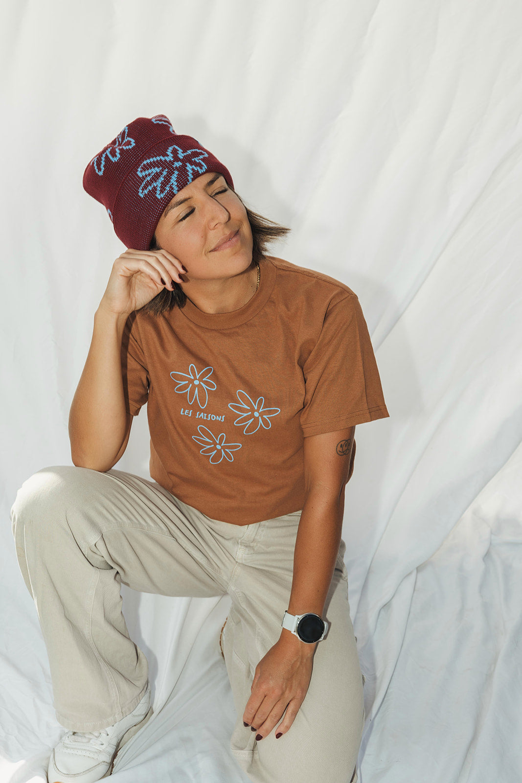Woman sat down with light brown t-shirt wearing dark red beanie with light blue flower pattern