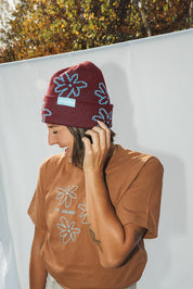 Woman with light brown t-shirt wearing dark red beanie with light blue flower pattern 